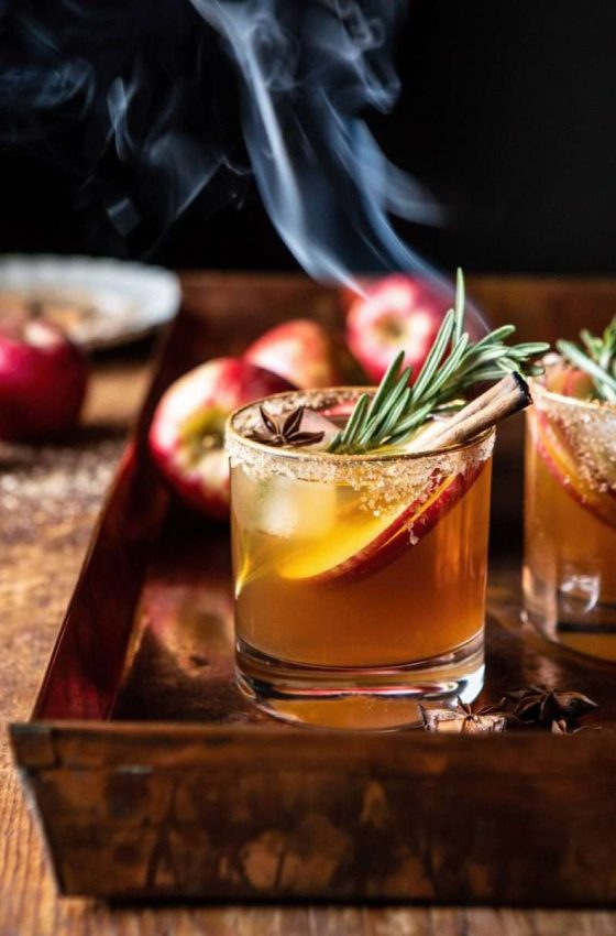 a tray glasses rimmed with sugar holding apple-cider margaritas with apple slices, rosemary, and a smoking cinnamon stick inside glass and apples in the background