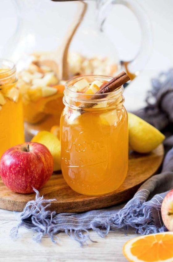 round wooden board with mason jars filled with a sparkling drink, diced apples, and a cinnamon stick