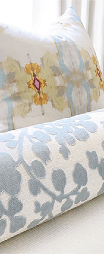 top of bed with 2 euro shams in impressionist print and blue and white cut velvet bolster with lamp in background