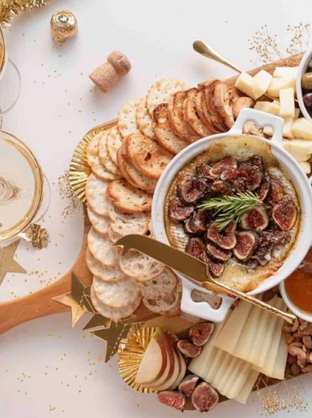 a wooden board showcases baked brie in a crock with sliced figs on top, fresh slices of apples and figs and cheese, crackers & crostini, ramekins of honey and olives