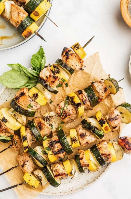 a platter with 4 long stainless steel skewers with chicken, mushrooms, zucchini and basil
