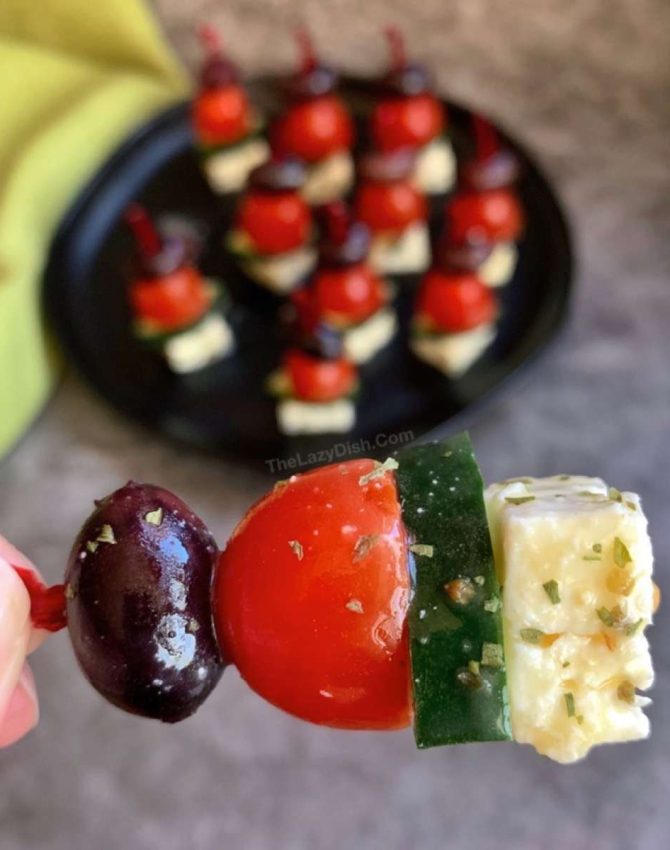 a skewer with a block of feta cheese, a grape tomato, a cucumber round and a black olive dripping with herb vinaigrette