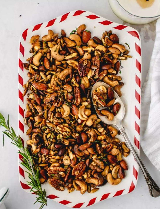 a red and white platter of roasted mixed nuts with a long sprig of rosemary