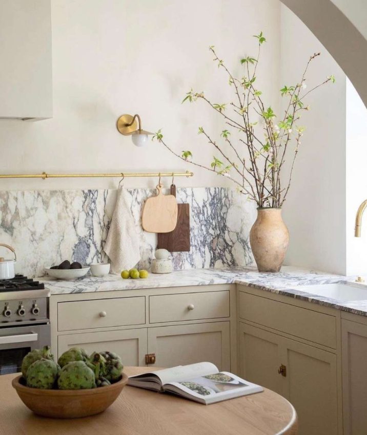 white walls, pale. mushroom cabinetry, heavy-veined marble counters and backsplash, styled with woods and textured ceramics, plants & artichokes