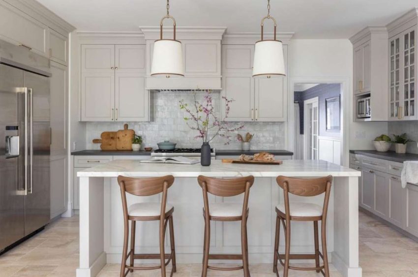 white walls, stone flooring set in French pattern, creamy cabinetry, off-white variegated zellige backsplash, quartz island countertop, soapstone countertops on the perimeter, brass, walnut & linen pendants over island, walnut counter stools, styled with textured ceramics and wooden cutting boards