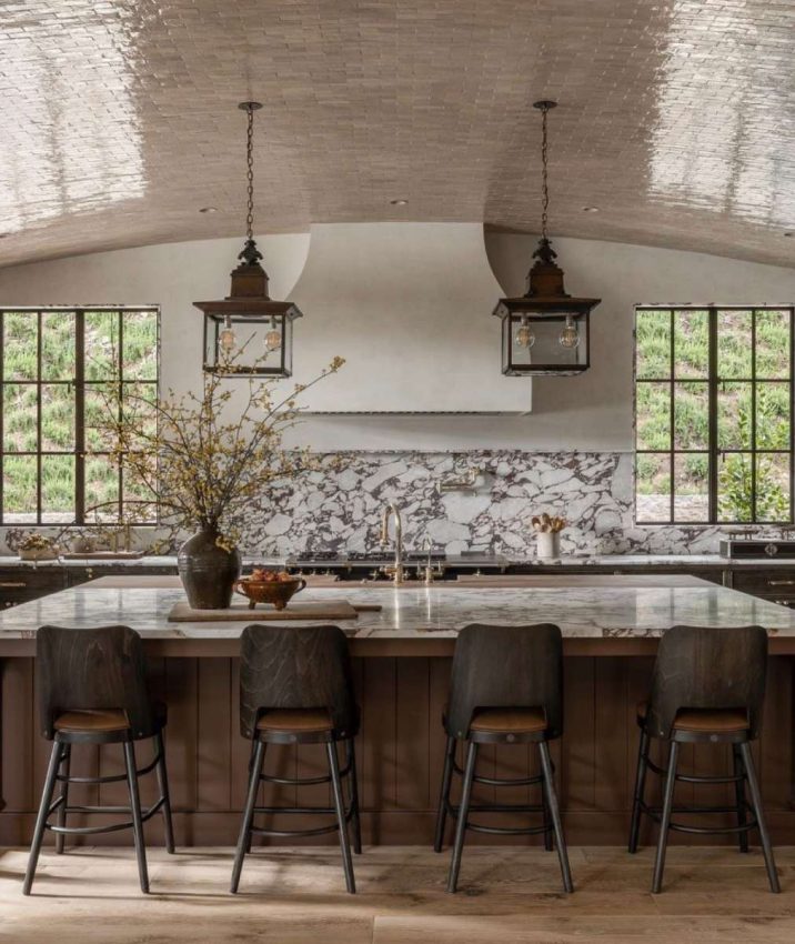 off-white plaster walls and hood vent, arched ceiling tiled with variegated glossy subway tile, med-brown wood cabinetry and island, heavy-veined marble countertops and backsplash with shelf, dark wood & leather counter stools, 2 large bronze lanterns over island, styling with wood, textured pottery, and large branches