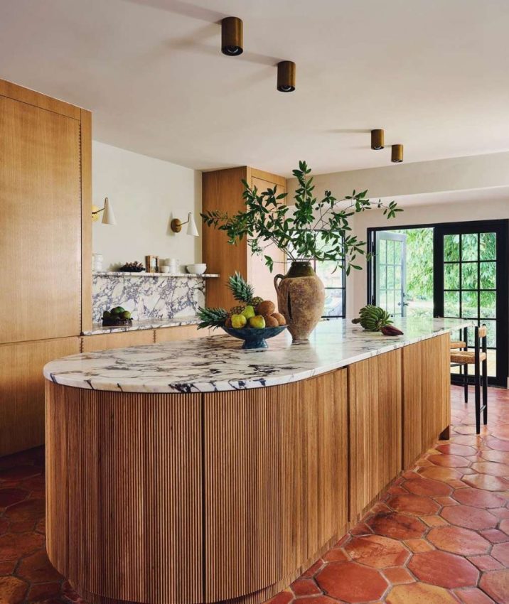 original saltillo tile flooring, light wood cabinetry, XL oval island with reeded wood and heavy-veined marble counter, marble backsplash with shelf, black-paned window and french doors, styling with white dishes, textured pottery and greenery