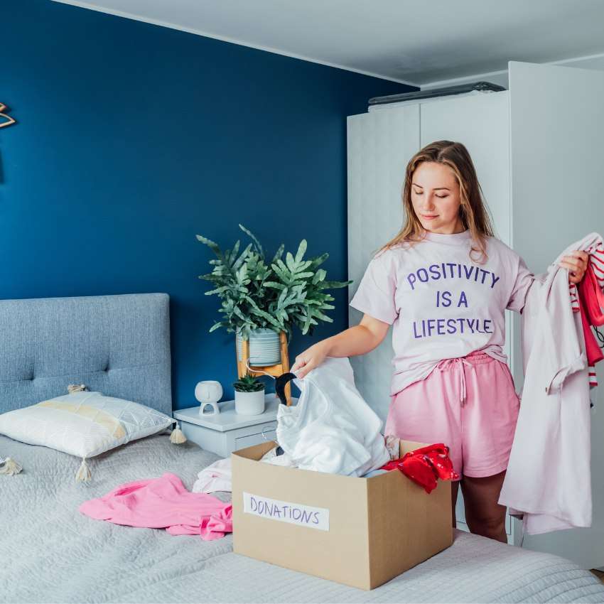 She's got a goal to declutter your home - a woman sorting clothes to put into a Donations box in a pretty bedroom
